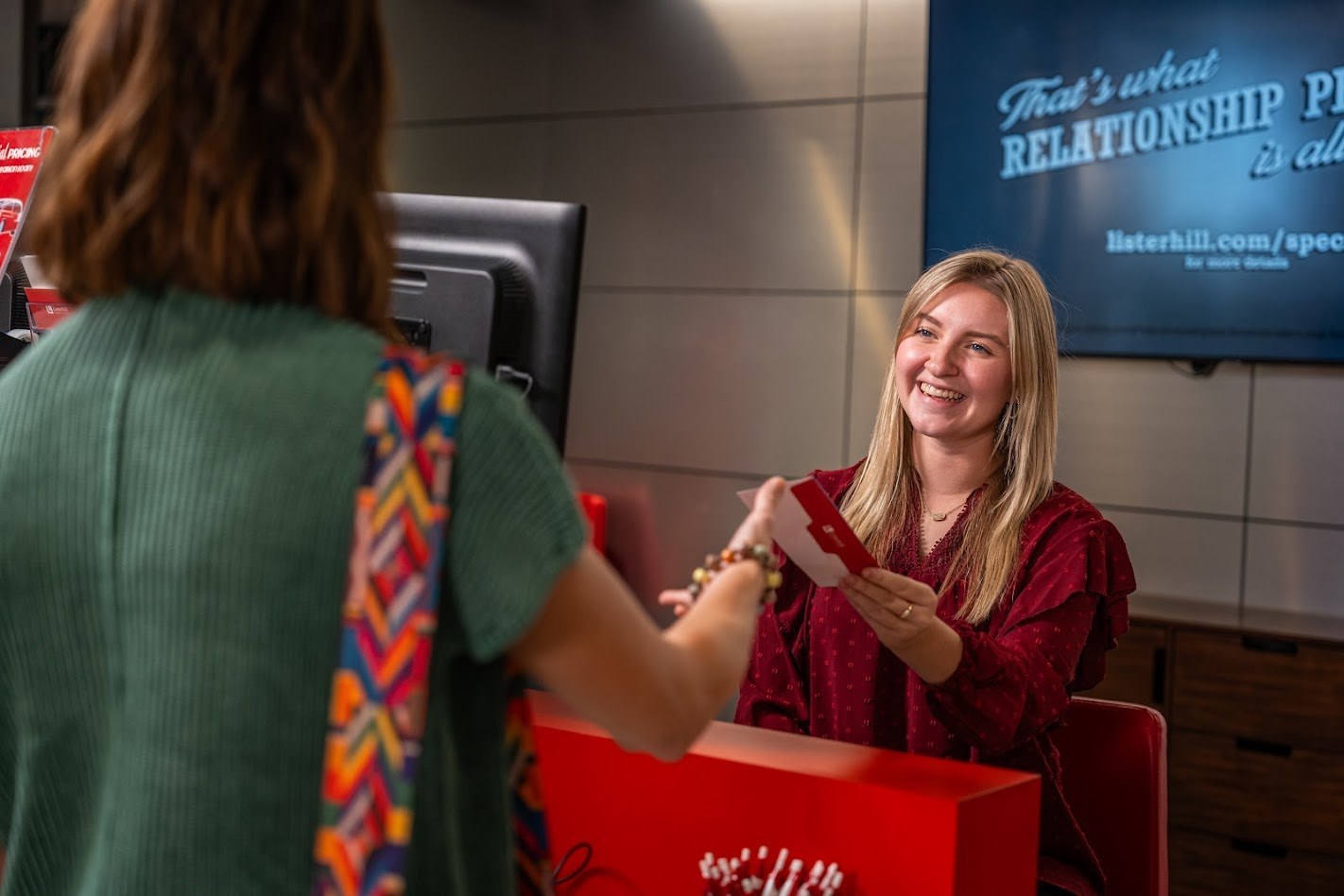 A team member assists a customer at a branch location, handing materials across the service counter during an in-person banking interaction.