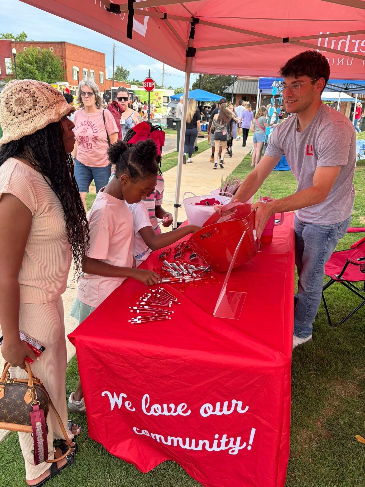 A Listerhill team members engages with families at a community event booth, distributing promotional items and information under a branded tent at an outdoor gathering.
