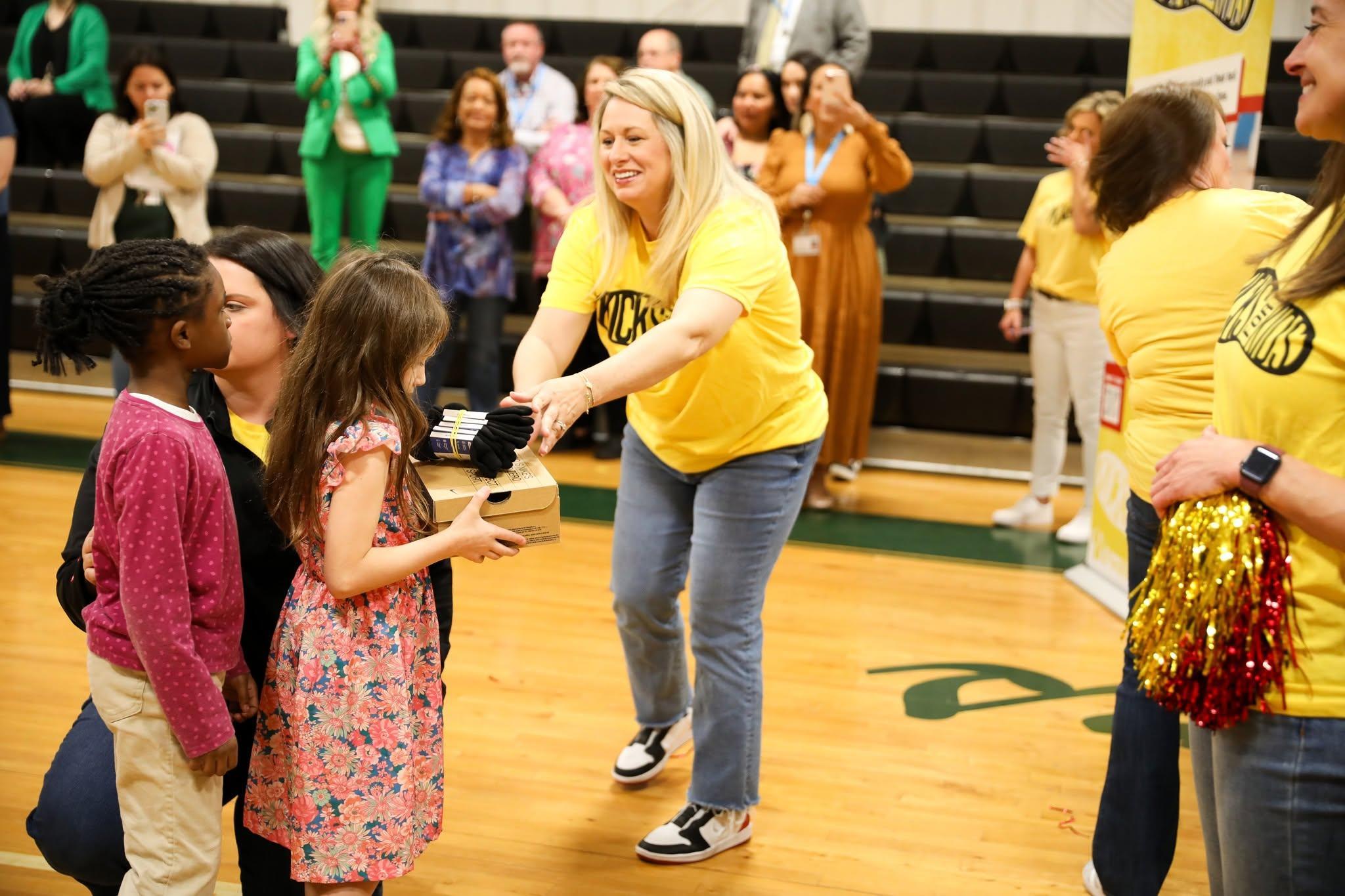 A team member presents a gift to children during a Kicks for Kids Shoe Drop held in a school gymnasium, with volunteers and attendees gathered in the background.
